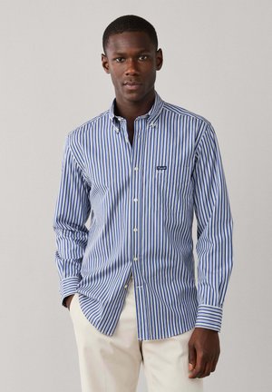 Man wearing a blue and white vertical striped button-up shirt with beige pants, standing against a plain light background.