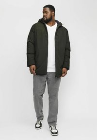 Donkergroen gewatteerde jas met een capuchon, voorzien van een ritssluiting zak, gedragen over een wit T-shirt, in combinatie met grijze spijkerbroek en zwarte sneakers.