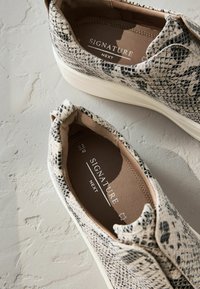 Chaussures à enfiler avec un motif serpent texturé en noir et crème, présentant un intérieur beige et une semelle en caoutchouc blanche. Marquage sur les semelles intérieures.
