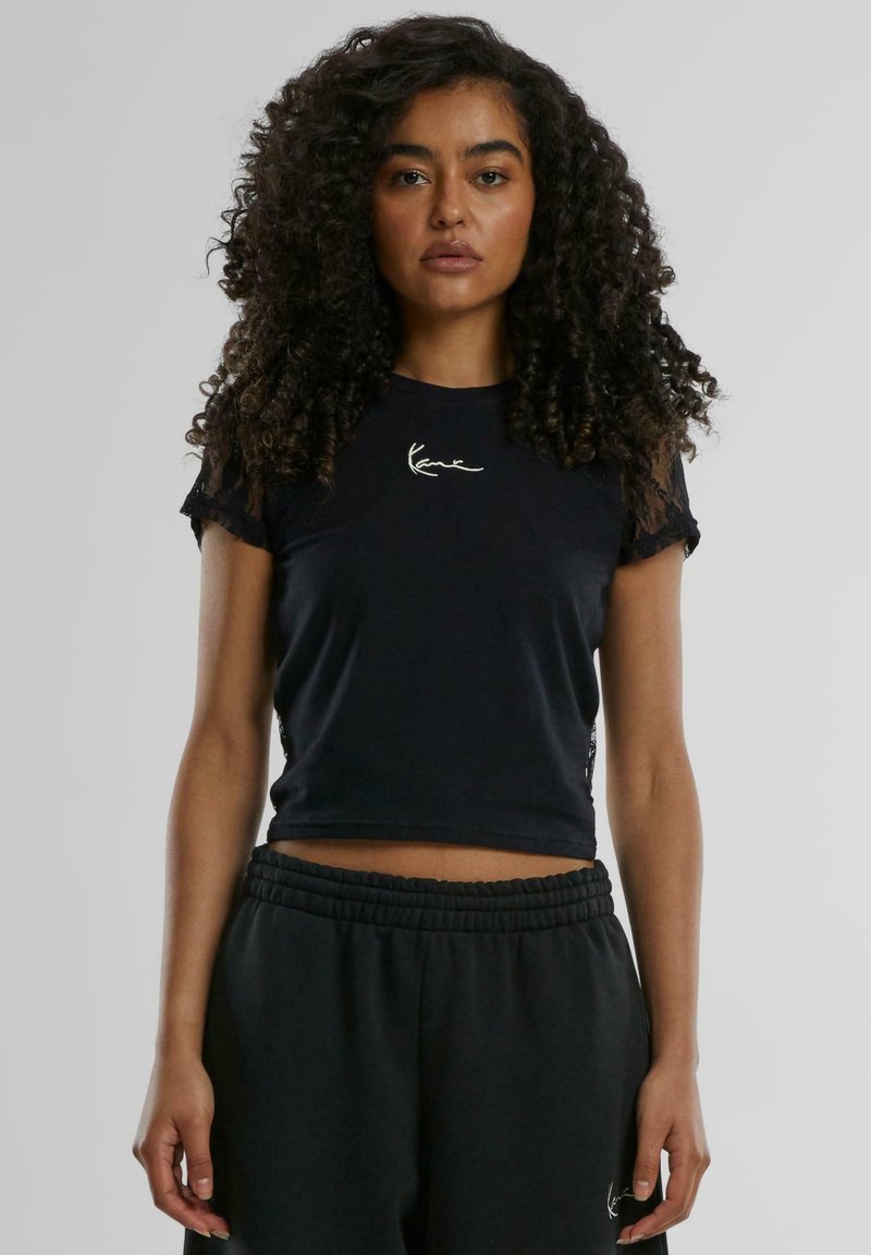 Svart cropped t-skjorte med en tettsittende design, korte blonde ermer og en sølvlogo på brystet. Kombinert med svarte elastiske joggebukser.