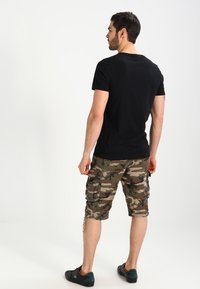 Schwarzes T-Shirt mit kurzen Ärmeln, kombiniert mit camo Cargo-Shorts, die über Taschen und einen knielangen Schnitt verfügen. Modell trägt dunkles Schuhwerk.