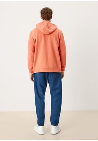 Oranger Kapuzenpullover mit lockerem Schnitt, kombiniert mit blauer Jeanshose. Der Stoff wirkt weich, und das Outfit hat gerippte Bündchen an Ärmeln und Saum.