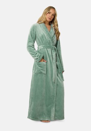Femme portant une robe verte douce longue jusqu'au sol avec une ceinture nouée à la taille, debout avec une main dans une poche avant.