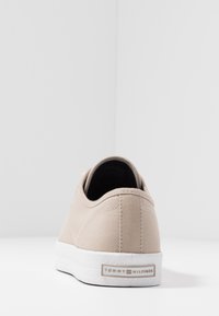 Beige Sneaker mit weißer Sohle und Tommy Hilfiger Logo an der Ferse, von hinten auf einer weißen Oberfläche vor einfarbigem Hintergrund gezeigt.