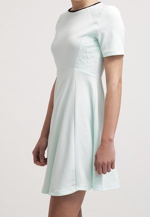 Femme portant une robe bleu clair texturée à manches courtes avec des panneaux en dentelle sur les côtés et une taille cintrée, debout sur un fond uni.