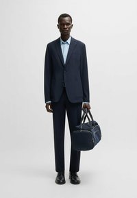 Costume bleu marine avec un blazer ajusté et un pantalon slim, associé à une chemise bleu clair. Tenant un sac de voyage bleu foncé, portant des chaussures noires.