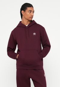 Egy férfi bordó Adidas kapucnis pulcsit és hozzá illő nadrágot visel, kezei a pulcsi zsebében, oldalra néz egy sima fehér háttér előtt.