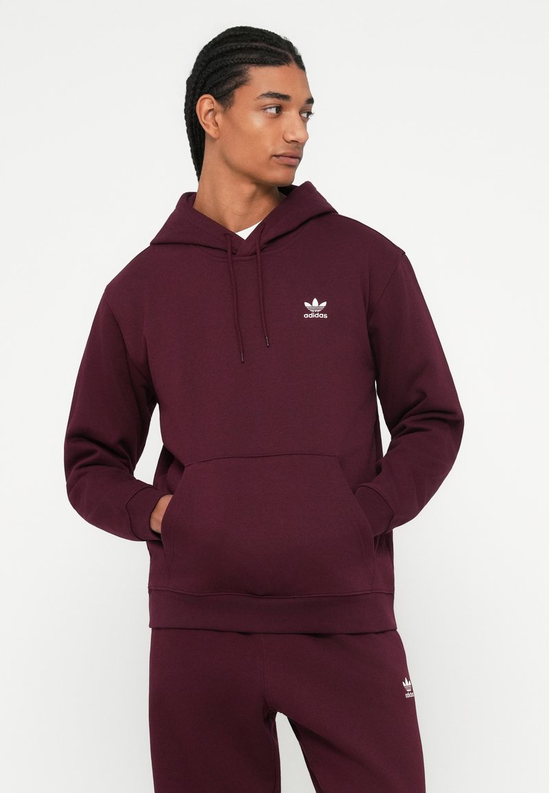 Mężczyzna w bordo bluzie Adidas i dopasowanych spodniach, z dłońmi w kieszeniach bluzy, spoglądający w bok na tle jednolitej białej ściany.