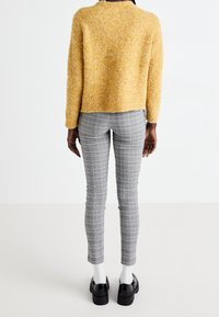 Persoon staat, draagt een donzige gele trui, grijs geruite slim-fit broek, witte sokken en zwarte platform schoenen tegen een eenvoudige witte achtergrond.