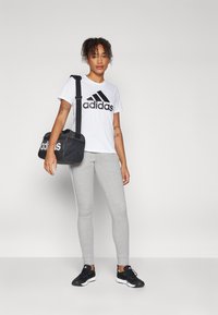 Biely bavlnený tričko s čiernym logom "adidas", šedé legíny, čierne športové topánky a čierna taška s bielym textom. Stojaci postoj na neutrálnom pozadí.
