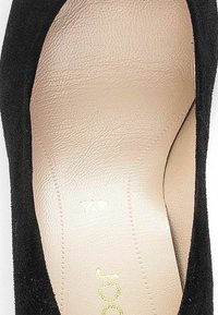 Escarpin noir en suede à talons hauts avec une semelle intérieure en cuir beige lisse. Présente une pointe arrondie et un design minimaliste, mettant en valeur des lignes épurées.