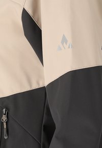 Gros plan sur la manche d'une veste beige et noire avec une fermeture éclair et un logo triangulaire réfléchissant sur le haut du bras.
