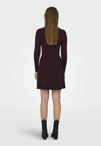 Femme aux longs cheveux portant une robe en maille côtelée violet foncé et des bottines à talons noires, vue de dos sur un fond clair uni.