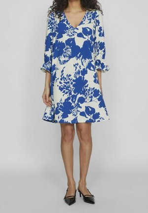 Vrouw draagt een witte jurk tot op de knie met blauw bloemenpatroon en driekwart mouwen met ruches, gecombineerd met zwarte pumps met een spits toelopende neus.