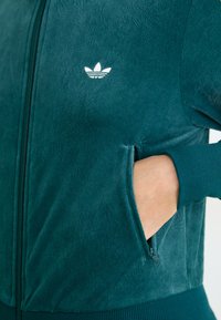 Mão inserida no bolso com fecho de correr de uma jaqueta de fleece Adidas em verde-azulado escuro, exibindo o logotipo no peito e o punho canelado na manga.