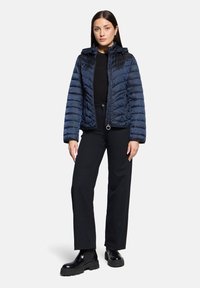 Doudoune bleu marine avec une silhouette ajustée, design matelassé vertical, col montant et poches zippées. Portée sur un haut et un pantalon noirs.