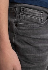 Jeans in denim grigio con una superficie testurizzata, cuciture in vista e dettagli in metallo nero sulla tasca frontale. Mano che riposa nelle vicinanze.