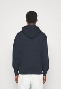 Osoba oblečená v tmavě modré mikině s kapucí a bílých kalhotách, zobrazená zezadu na jednobarevném světlém pozadí.