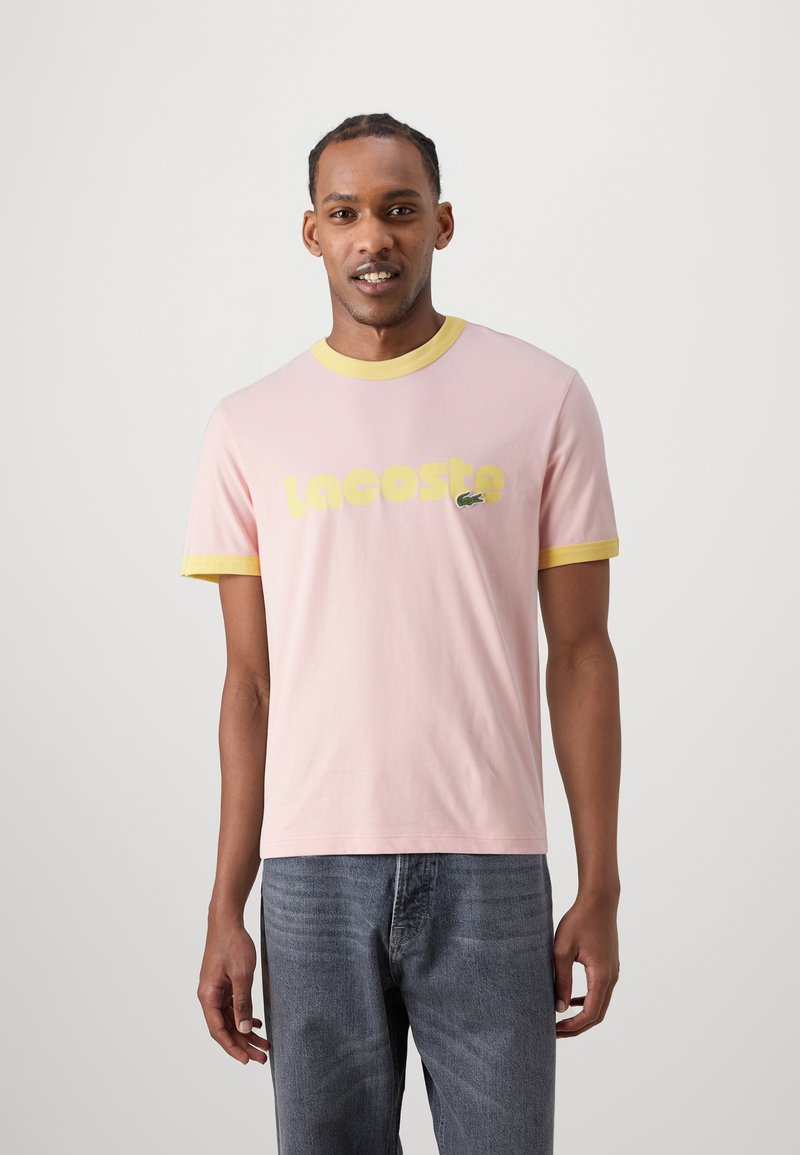 Camiseta rosa con cuello y mangas acanaladas amarillas, con el logo "Lacoste" en texto amarillo en negrita y un pequeño acento de cocodrilo verde.