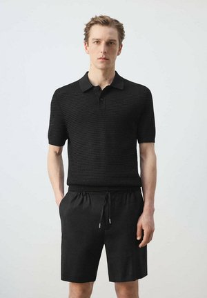 Joven de pie vestido con una camiseta polo negra texturizada de manga corta y pantalones cortos negros con cordón, sobre un fondo claro y liso.