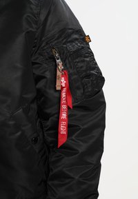 Veste bomber en nylon noir avec une poche zippée, une étiquette rouge "REMOVE BEFORE FLIGHT" et un tissu-texturé avec une finition lisse.