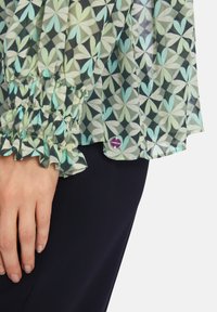 Durchsichtige Bluse mit einem grün-schwarzen Blumenmuster, gerafften Manschetten und einem lila Logoetikett am Saum, kombiniert mit dunklem Stoff darunter.
