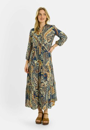 Mujer con vestido de manga larga hasta los tobillos con estampados de cachemira en azul, marrón y crema, combinados con sandalias marrones de plataforma.