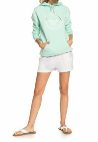 Mátově zelená mikina s zlatým logem, doplněná světle tie-dye šortkami. Obsahuje přední kapsy a bílé žabky. Neformální letní outfit.
