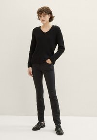 Juodas megztinis su V formos iškirpte ir tekstūruotu raštu, derinamas su juodomis siaurėjančiomis kelnėmis ir juodais batais. Modelis stovi su rankomis kišenėse.