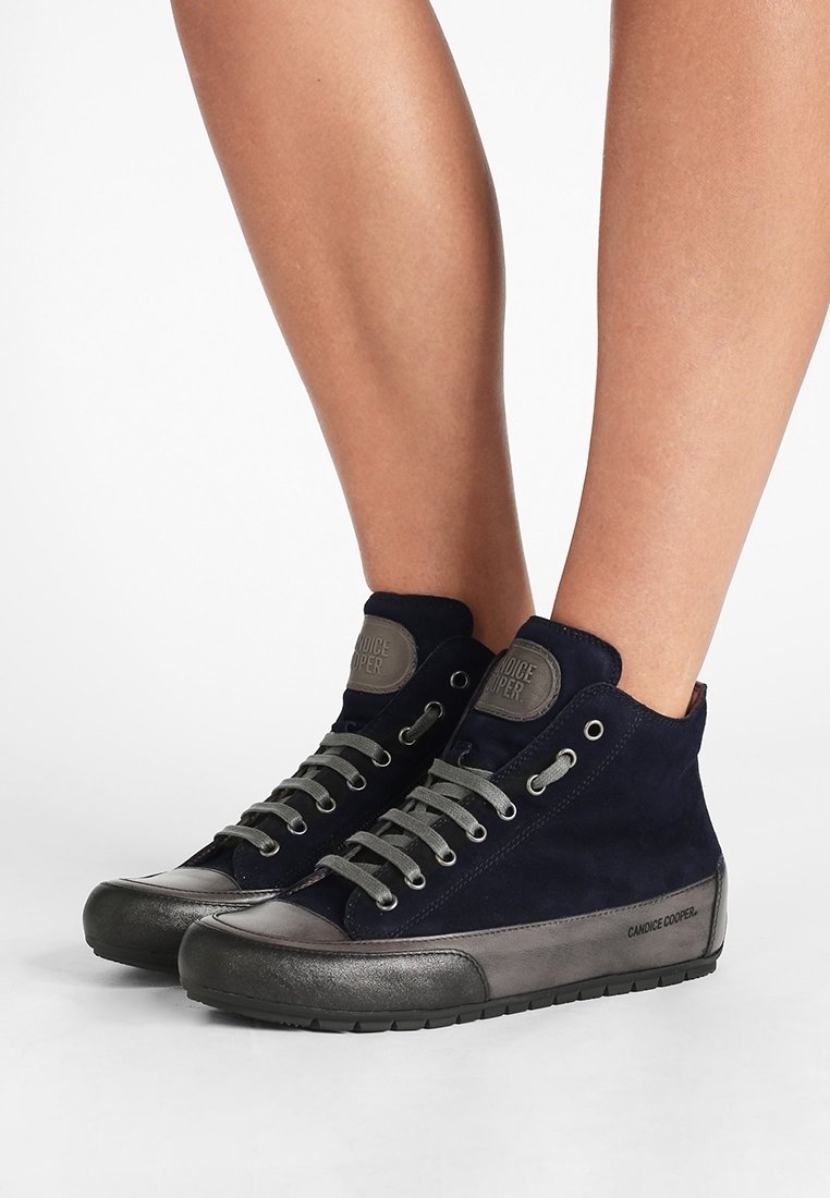 Personne portant des baskets montantes noires et grises avec des lacets gris, debout sur un sol blanc contre un fond blanc uni.