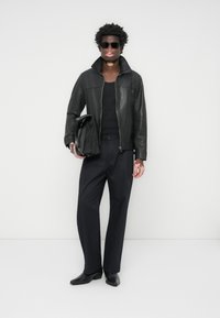 Black leather jacket with a zip closure, paired with a black tank top and tailored trousers. Holds a black bag, wearing sunglasses.