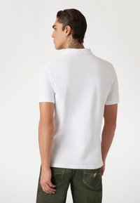 Weißes Poloshirt mit kurzen Ärmeln und Kragen, getragen mit grünen Hosen. Glatte Textur, taillierte Passform, keine sichtbaren Muster oder Logos.