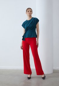 Teal short-sleeved top with a twisted front design paired with bright red wide-leg trousers. Black high heels complement the outfit.