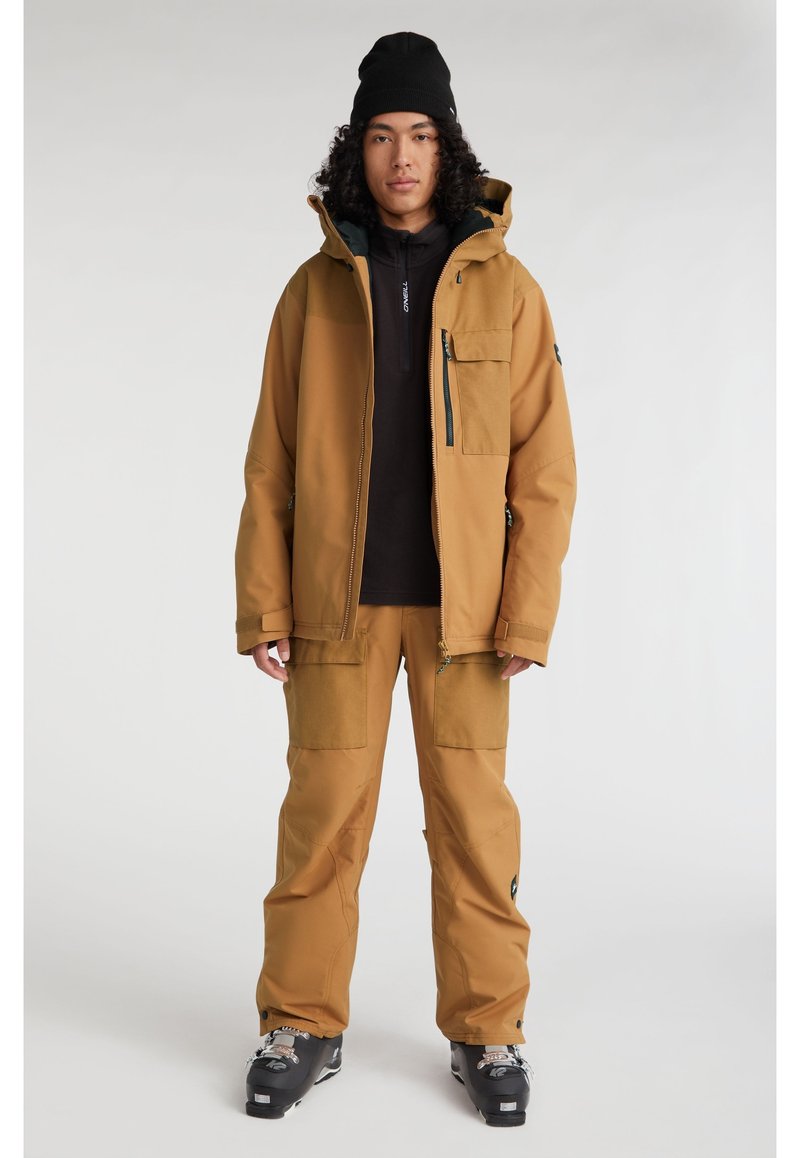 Braune wasserdichte Jacke mit Kapuze, Reißverschlusstaschen und passenden Cargo-Hosen. Ein schwarzes Basislayer und Schneestiefel runden das Outfit ab.