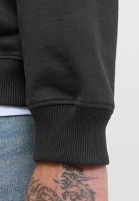 Mørkegrå sweatshirt-stof med ribbet detalje på ærmerne; synlig arm-tatovering; teksturen virker blød og let strækbar.