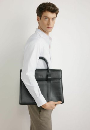 Młody mężczyzna w białej koszuli trzyma czarną skórzaną teczkę na jasnym, jednolitym tle, patrząc w stronę kamery.