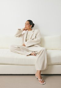 Blazer oversize beige et pantalon large associés à un pull léger. Le mannequin porte des sandales à talons blanches, assise sur un canapé de couleur crème.