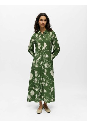 Femme portant une robe verte à manches longues boutonnée avec des motifs floraux blancs, debout, les mains derrière le dos et des ballerines beiges.