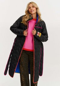 Čierny prešívaný dlhý kabát s jasne ružovými a žltými akcentmi, so zapínaním na gombíky a modrou podšívkou, nosený cez ružový sveter a vzorované nohavice.