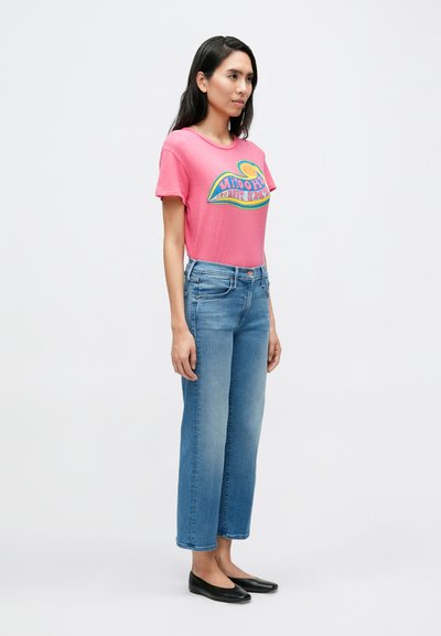 Roza grafička majica s šarenim dizajnom, uparena s ravnim trapericama svijetloplave boje. Crne ravne cipele upotpunjuju odjevnu kombinaciju.