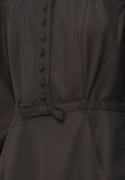 Tuvo plāns tumšas auduma kleitas, kas rotāta ar auduma pogām un plānu sasietu jostu viduklī.