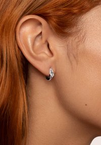 Orecchino a cerchio in argento con un accento a forma di cuore e una pietra preziosa trasparente. Superficie liscia e lucida con un design elegante e moderno.