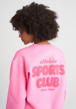 Kvinne med krøllete hår og solbriller iført en lys rosa hettegenser med teksten "ellesse SPORTS CLUB siden 1959" på baksiden.