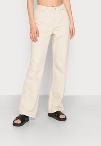 Jeans largos bege feitos de algodão, apresentam um design clássico de cinco bolsos e costuras laranja discretas. Combinados com sandálias pretas de tiras cruzadas.