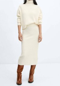 Pull en tricot côtelé crème avec un col montant, associé à une jupe midi côtelée assortie. Des bottes en cuir marron montant jusqu'aux genoux complètent l'ensemble.