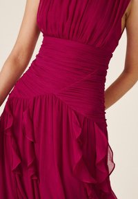 Vestido rosa intenso sin mangas con cintura fruncida y detalles de volantes en cascada en la falda, mostrado en una persona desde los hombros hasta las caderas.