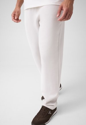 Homme portant un pantalon de survêtement blanc et des baskets marron foncé à semelles blanches, debout avec les bras détendus sur les côtés, fond neutre.