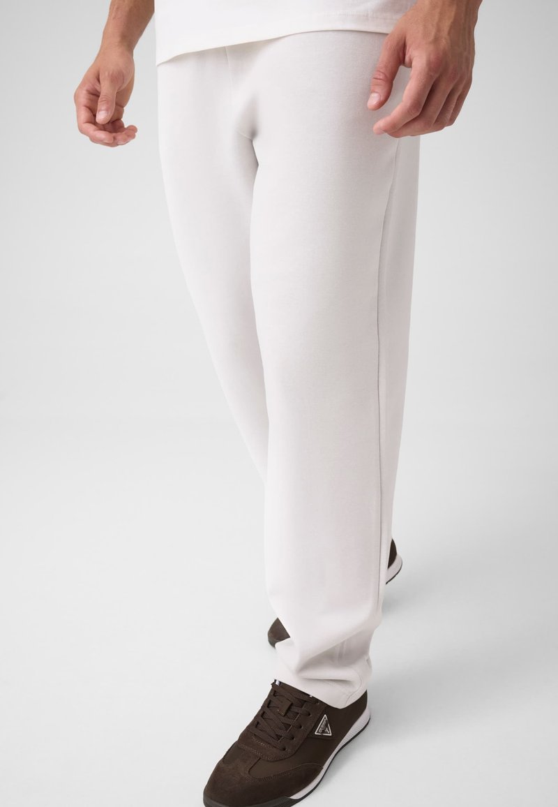 Homme portant un pantalon de survêtement blanc et des baskets marron foncé à semelles blanches, debout avec les bras détendus sur les côtés, fond neutre.