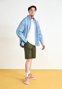 Cămașă din denim de culoare albastru deschis, cu nasturi, peste un tricou alb, pantaloni scurți de culoare verde măsliniu și adidași roz cu șosete albe, toate pe un podea galbenă.