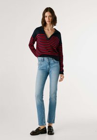 Nööriline villane sviiter tumesinisest ja punasest värvist, V-kaelusega ja soonikkudega varrukatega, koos helesiniste teksade ja mustade studskingadega.